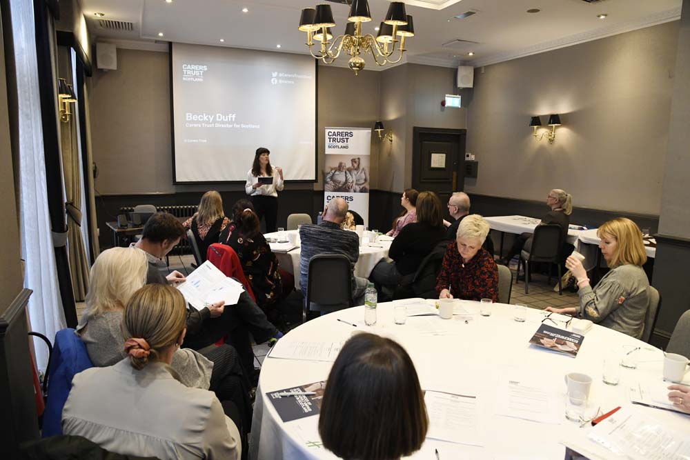 Becky Duff hosting a presentation to group of carers sitting around round tables.